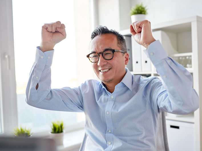 Man sitting at desk smiling with arms raised in celebration